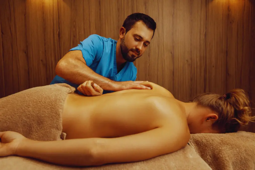 A woman receiving a back massage on a massage table. She is lying face down with her eyes closed, and a man in a blue shirt is massaging her back. The focus is on the woman's back and the masseur's hands. The background is a wooden wall with soft lighting, creating a relaxing atmosphere. The lighting is warm and focused, highlighting the details of the massage.
