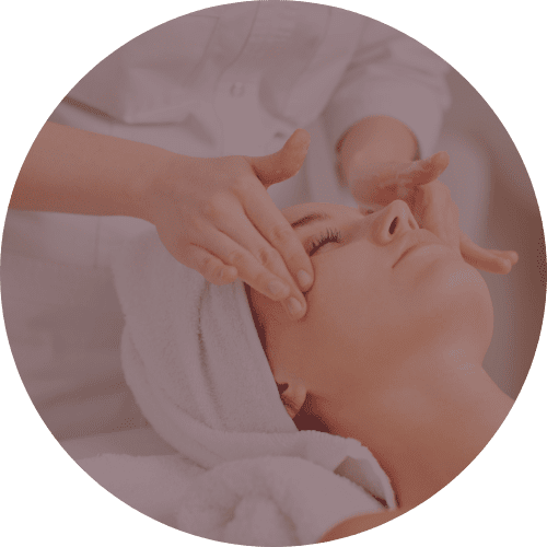 A circular close-up image of a woman receiving a facial massage. Her eyes are closed, and a towel is wrapped around her hair. A person in a white uniform is gently massaging her face with their hands. The image is cropped in a circle with a gray background. The lighting is soft, focusing on the woman's face and the hands providing the massage.
