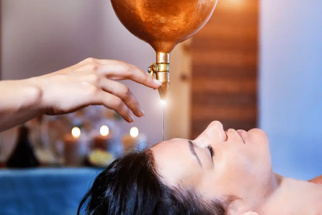 A close-up shot of a woman receiving a head massage. A golden pot hangs above her head, dripping a thin stream of oil onto her forehead. A hand gently guides the oil flow. The woman's eyes are closed, indicating relaxation. The background is blurred, with soft lighting and a hint of candles, creating a serene atmosphere.