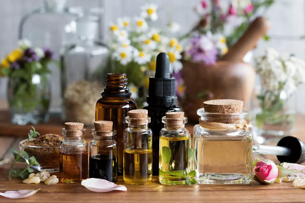 different oil for head massage A still life image featuring a collection of small glass bottles filled with various colored essential oils, arranged on a wooden surface. The bottles have cork stoppers and are of different sizes and shapes. In the background, there are more glass bottles, dried herbs, flowers in vases, and a wooden mortar and pestle, suggesting a natural or apothecary setting. The lighting is soft and natural, highlighting the textures of the wood and glass.