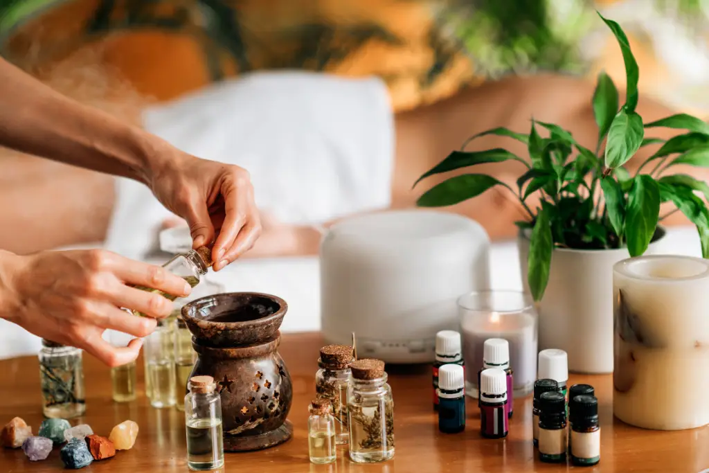 Close-up of hands pouring oil from a small bottle into a larger one, with other aromatherapy oils and massage setting visible.


