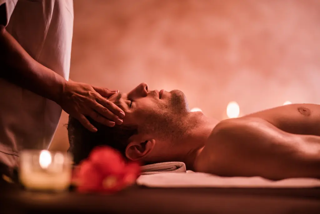 A man with dark hair lies on a massage table, eyes closed, receiving a head massage from a massage therapist. Candles and a red flower are visible in the foreground, creating a warm, relaxing atmosphere. The room is dimly lit with a reddish hue.