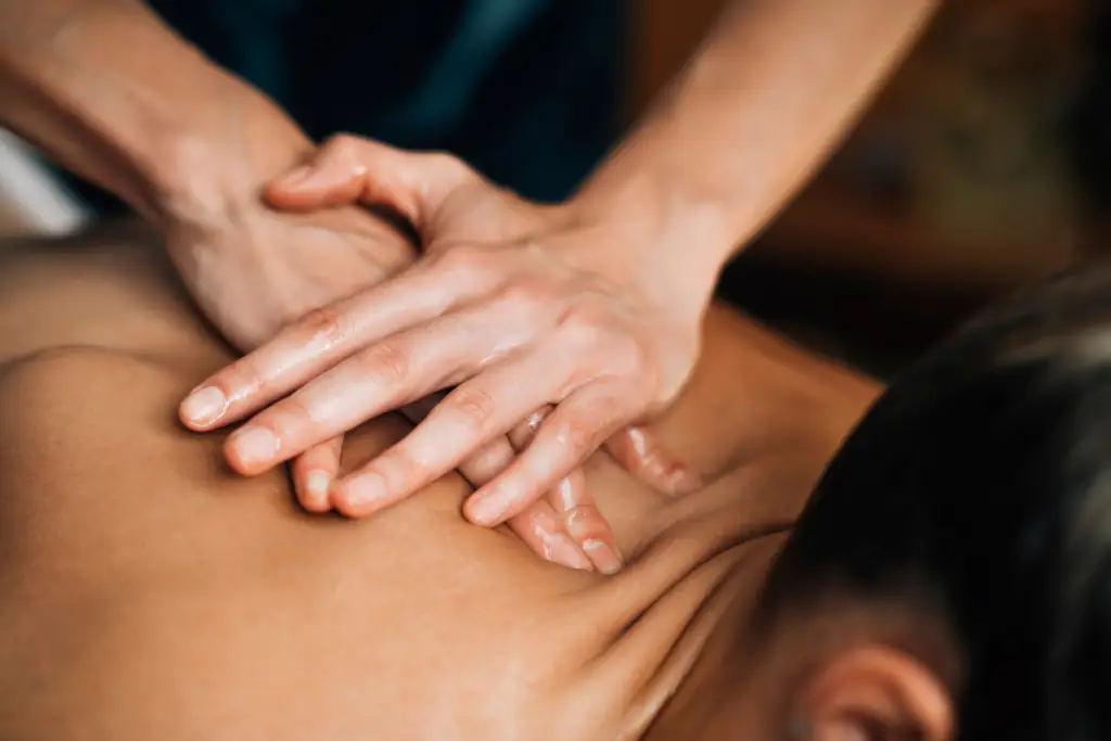 A close-up shot of a massage therapist's hands applying oil and massaging a person's bare back. The hands show technique and oil application, highlighting a massage for weight loss session. The person's head and shoulder are partially visible in the foreground.
