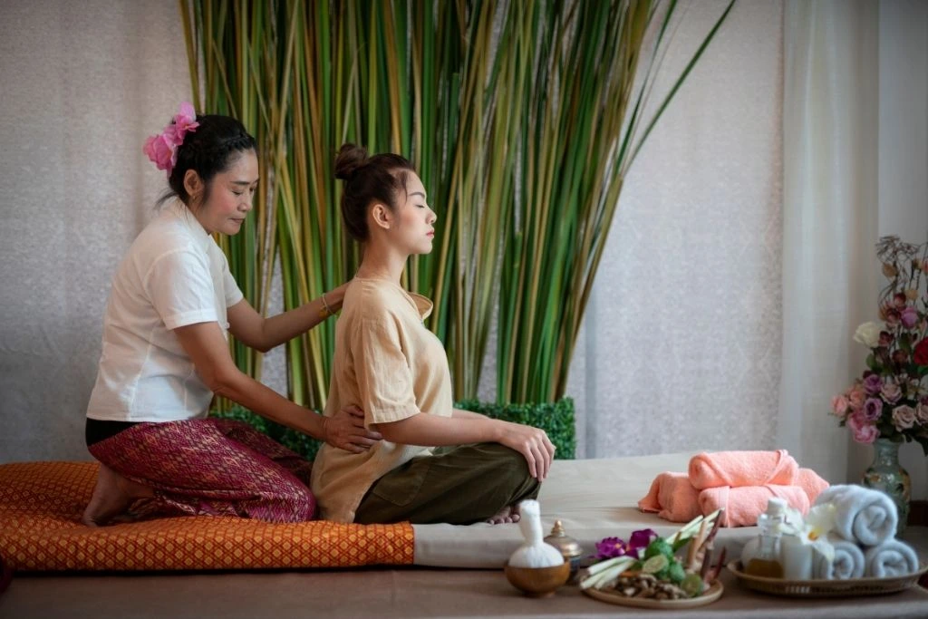 A massage therapist, who appears to be of Asian descent, kneels behind a seated client, applying pressure to their shoulders and back at Spa Restoration Center.